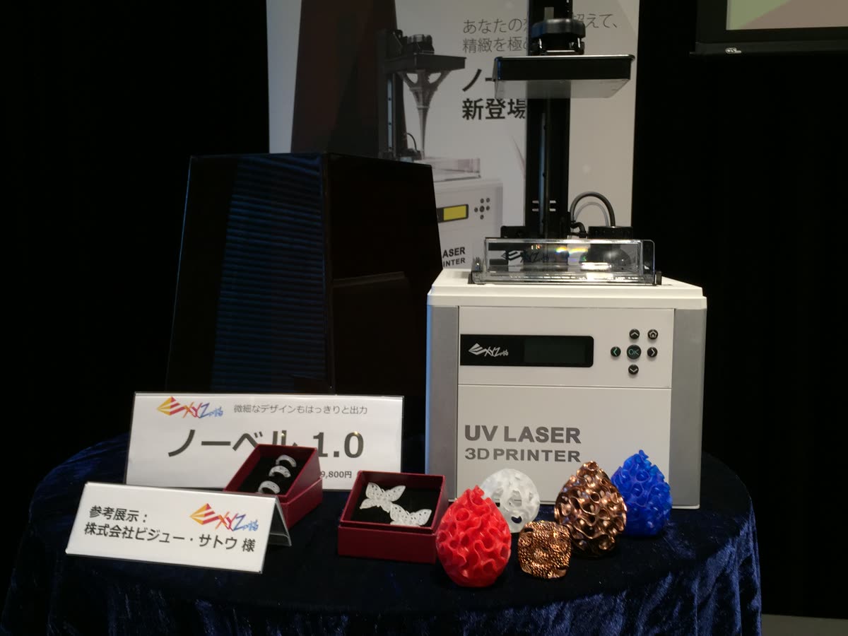 XYZプリンティング「ノーベル1.0」発表記者会見の展示台。「参考展示：株式会社ビジュー・サトウ様」の名札とともに、ノーベル1.0実機の隣に当社のジュエリー出力サンプル（リング・蝶のペンダント）が展示されている